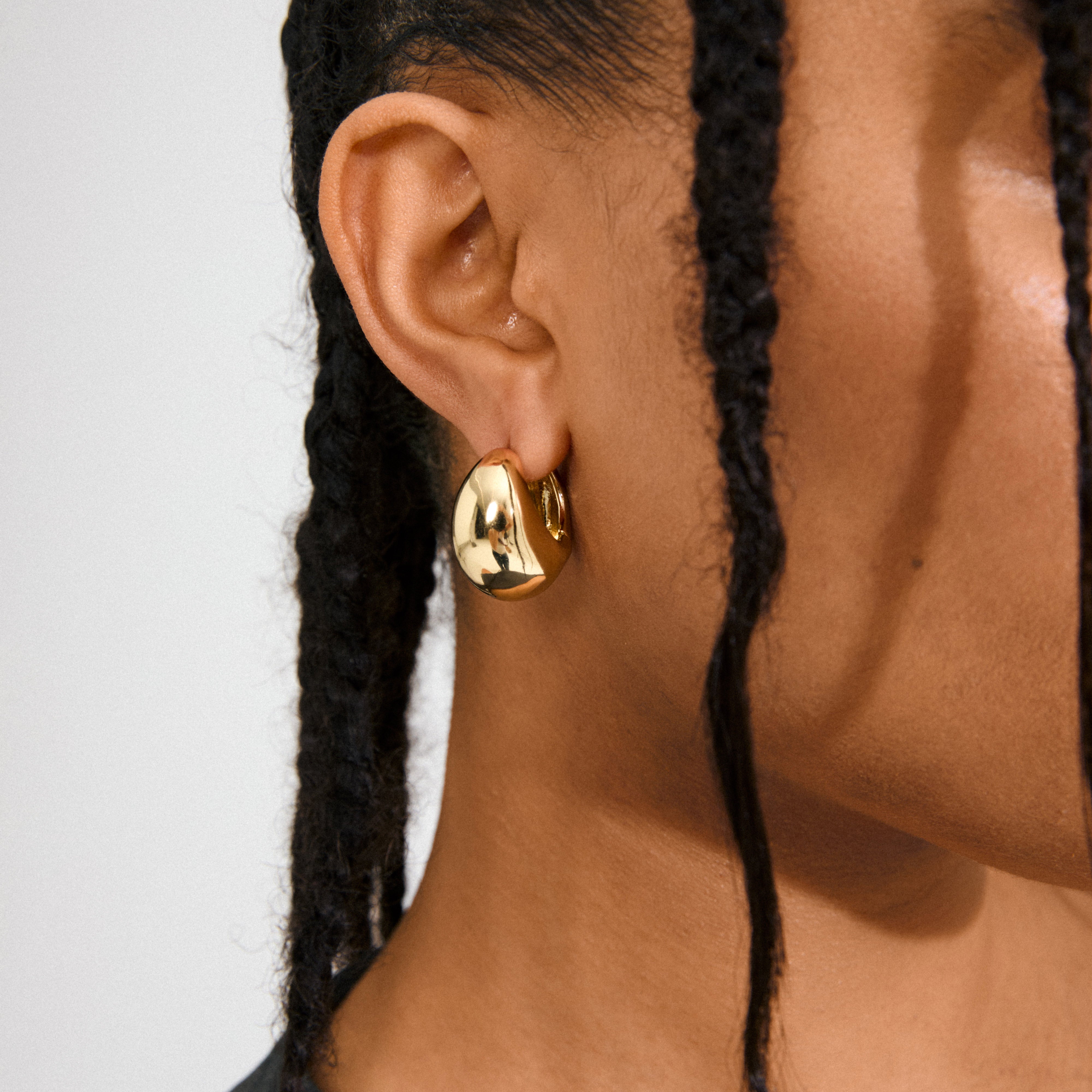 Close-up of a medium-dark skin ear wearing Pilgrim's FREE hoops 168一分钟极速赛车--|实时赛果直播视频 gold-plated, featuring a chunky, uneven sculptural design, framed by thin, dark braids.