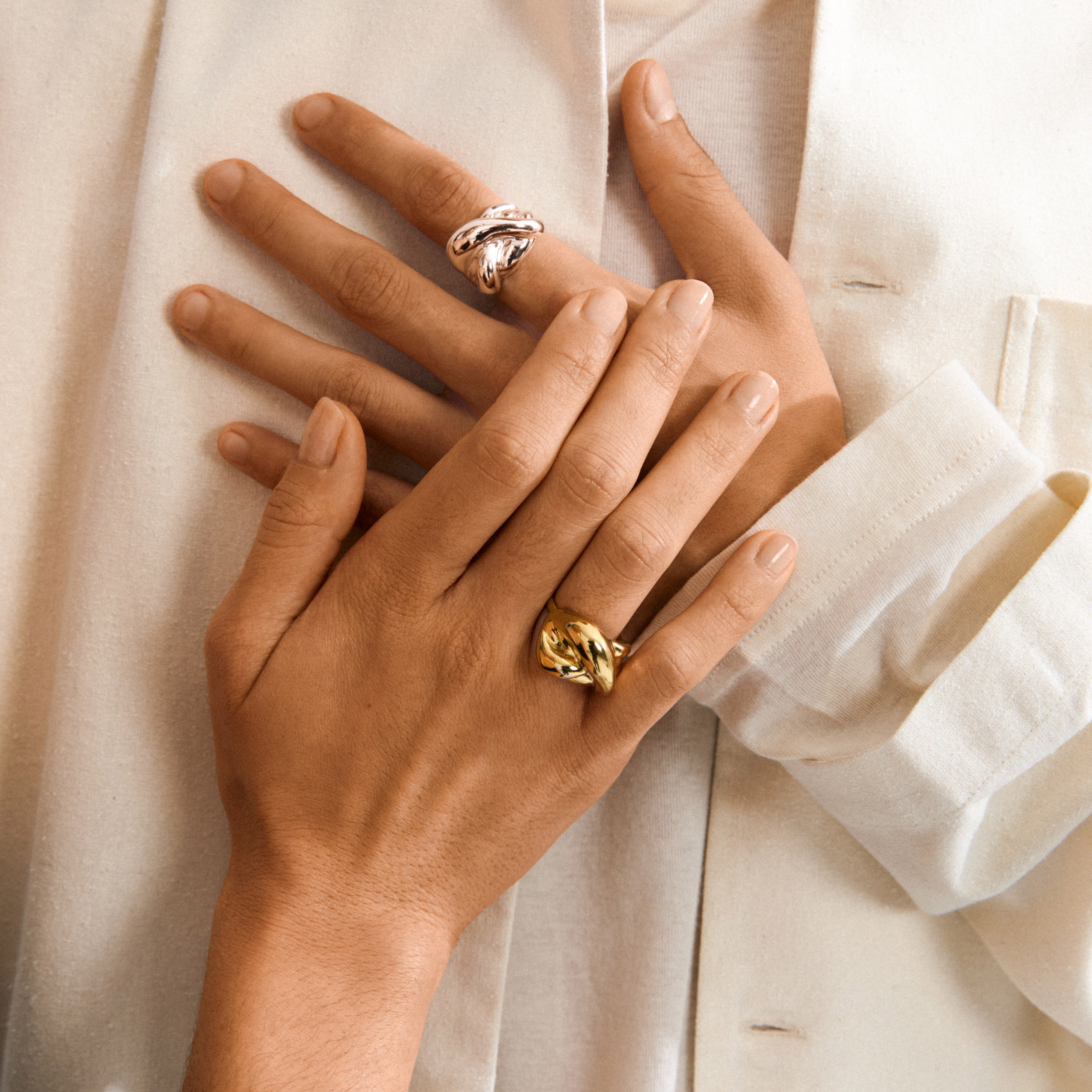 Hands overlapping on cream fabric, each adorned with the Pilgrim UNFOLD ring in gold-plated, showcasing a knotted, sculptural design with a luxurious finish.