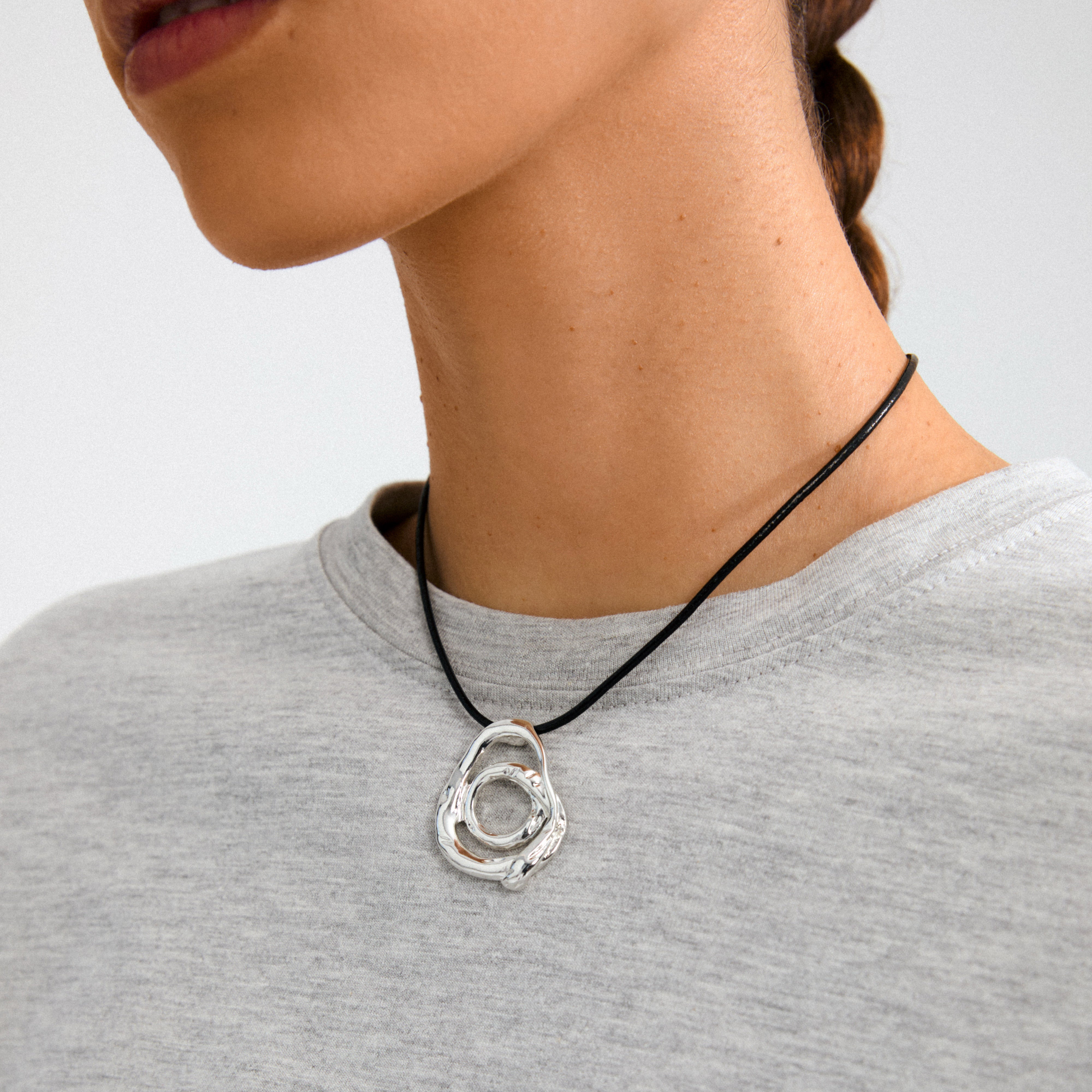 Close-up of a person wearing the UNFOLD necklace silver-plated by Pilgrim, showcasing its chunky, artistic silver pendant on a black cord against a light-gray top.