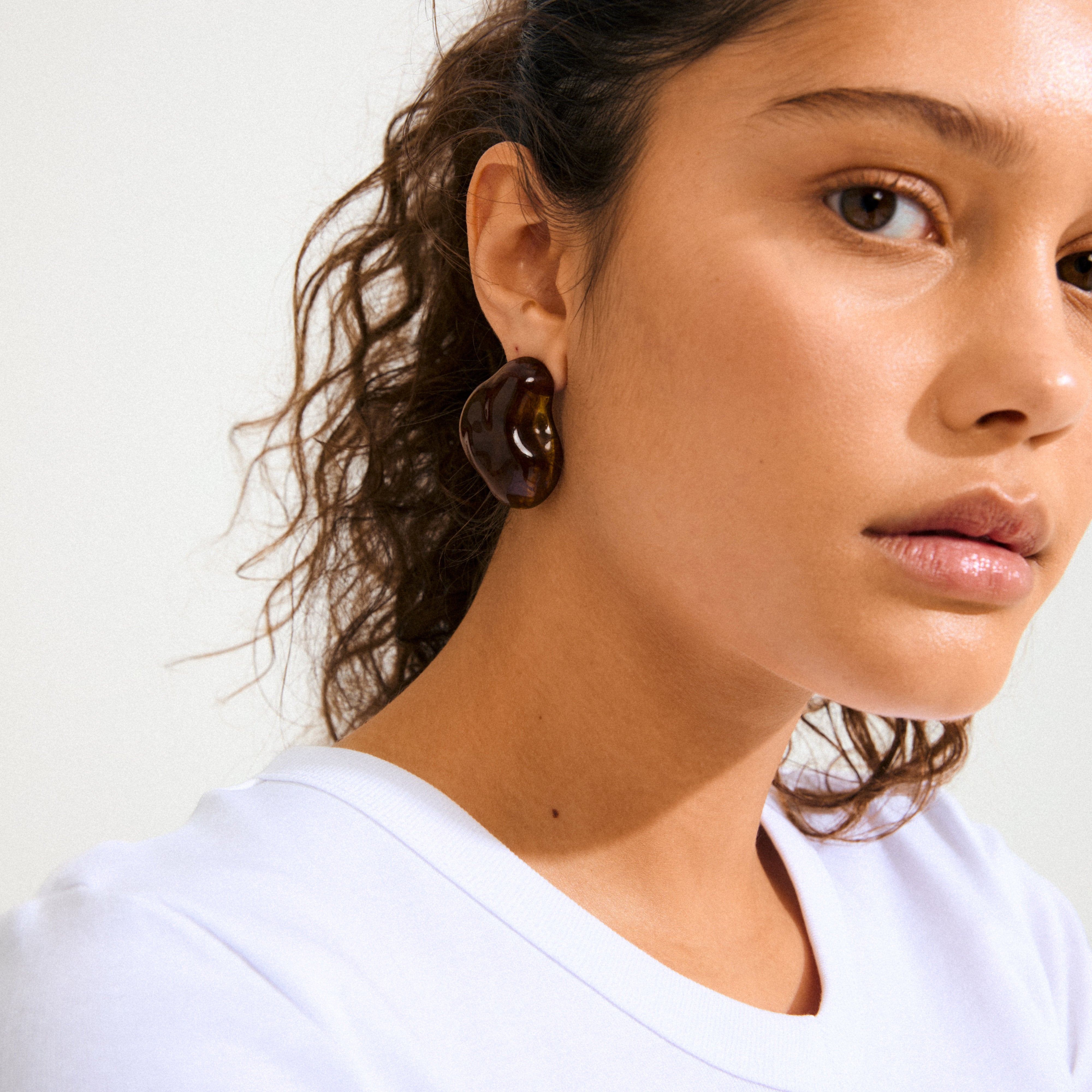 Young woman wears a chunky, dark brown, bean-shaped earring, the LIAN earrings gold-plated/brown from Pilgrim, showcasing a minimalist and bold style.