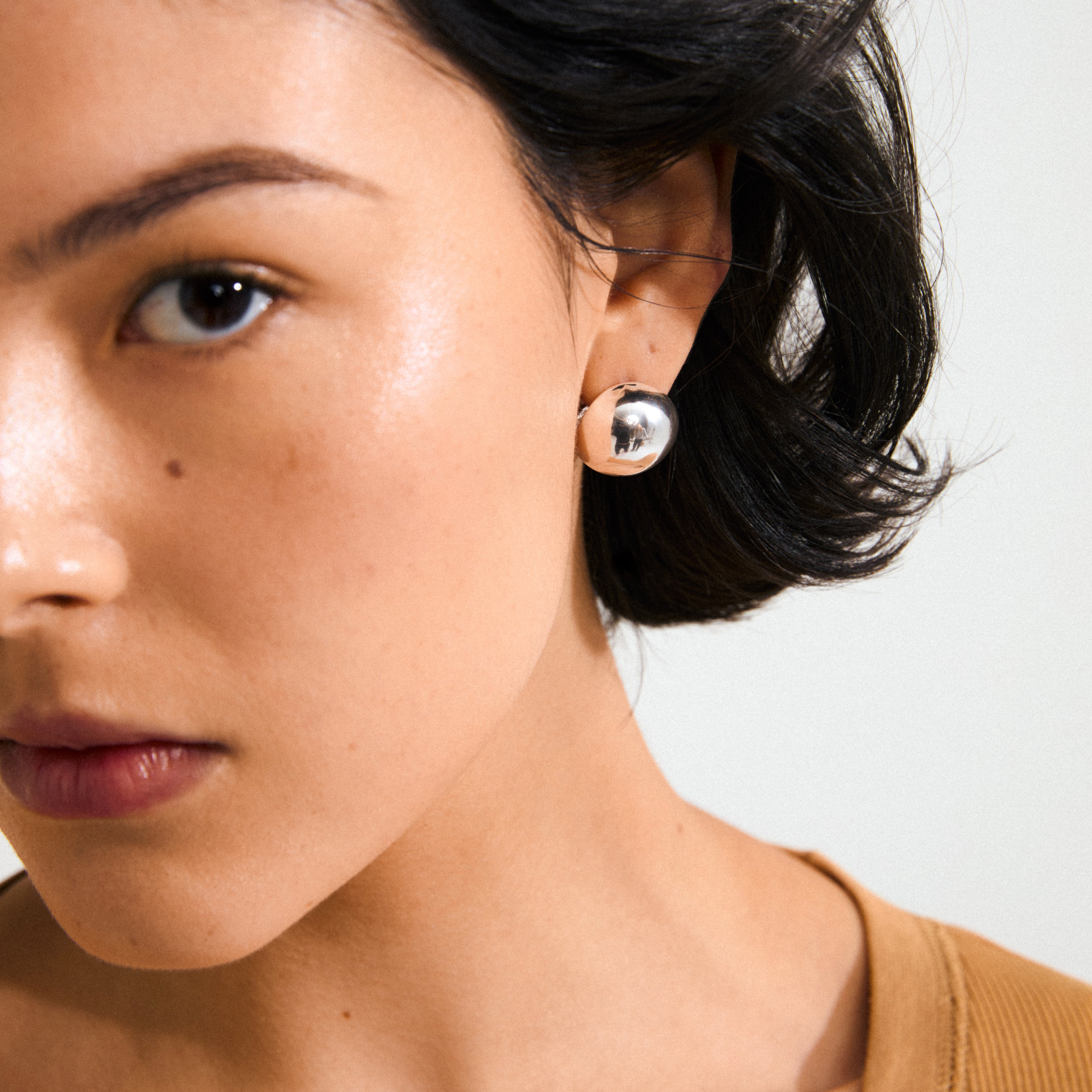 Close-up of the JACE silver-plated earrings by Pilgrim, showcasing a shiny, spherical design worn on a person with short, dark hair.